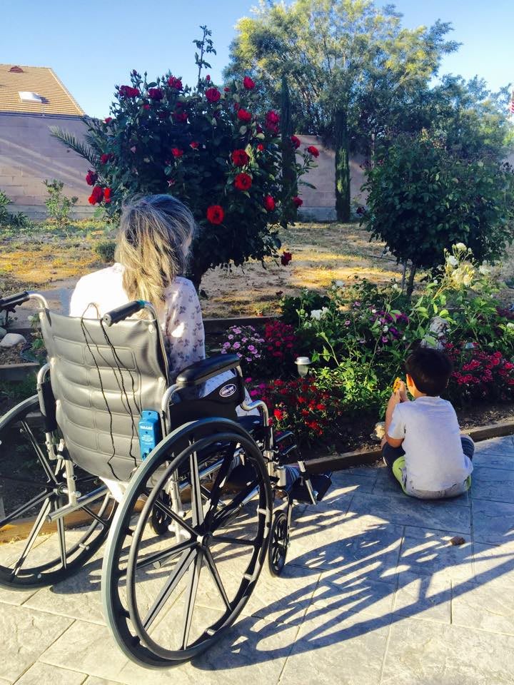 A person in a wheelchair and a child enjoying time together in a garden surrounded by flowers.