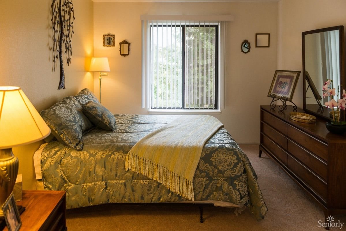 A cozy bedroom with a neatly made bed, warm lighting from a lamp, and natural light streaming through the window.