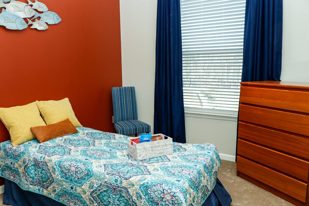A cozy bedroom with colorful decor, featuring a bed, chair, dresser, and a large window with curtains.