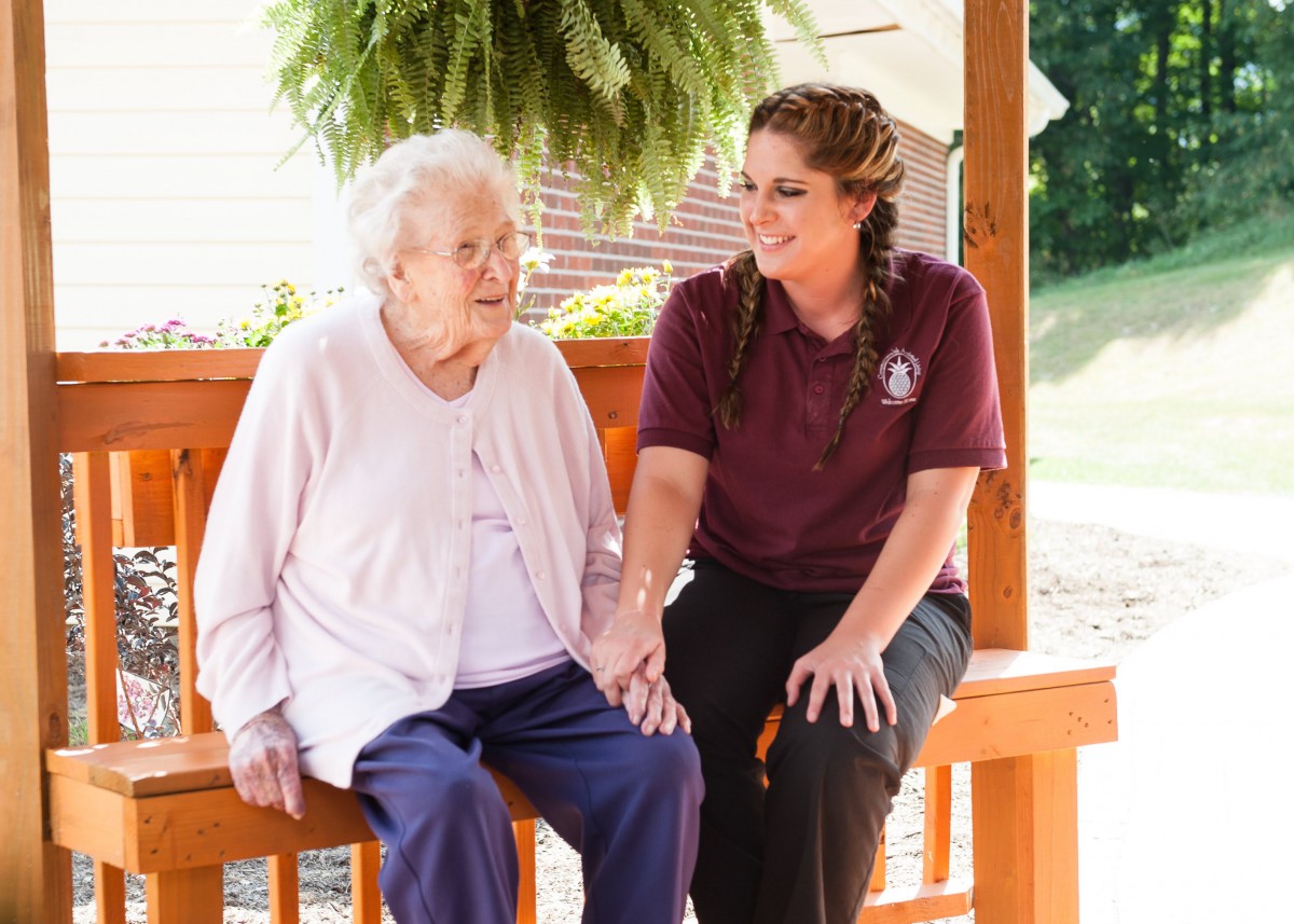 An elderly woman sitting with a caregiver on a bench, sharing a moment of companionship outdoors.