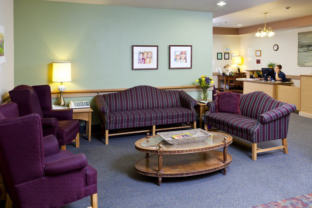 A cozy waiting room with purple sofas and chairs, a glass coffee table, soft lighting, and framed artwork on the walls.