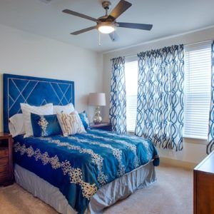 A cozy bedroom with blue bedding and patterned curtains, featuring a ceiling fan and stylish decor.