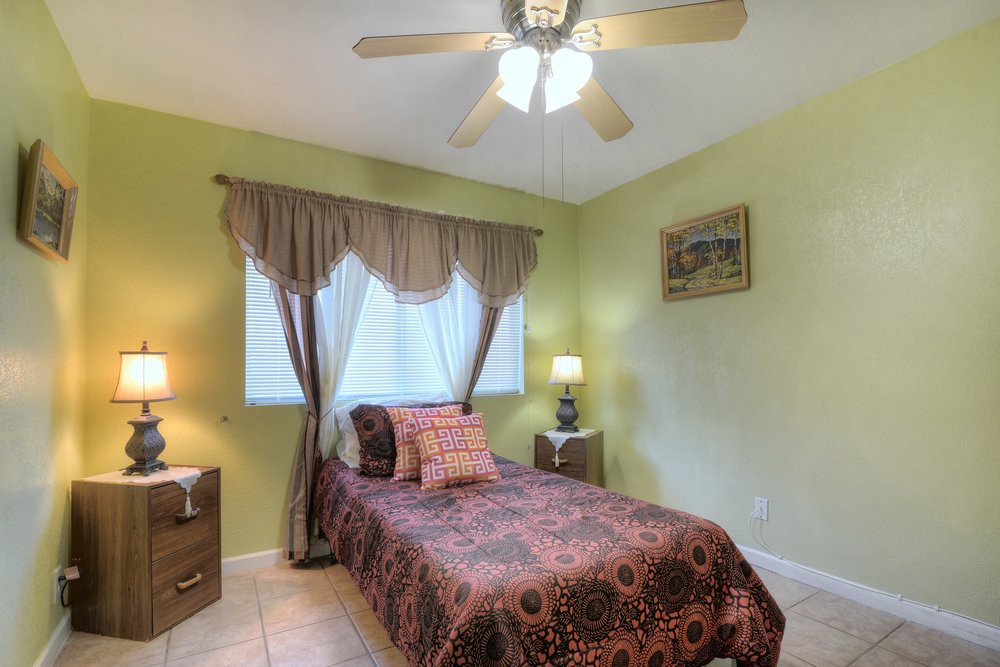 A cozy bedroom with a single bed featuring patterned bedding, flanked by nightstands with lamps, and a ceiling fan overhead.