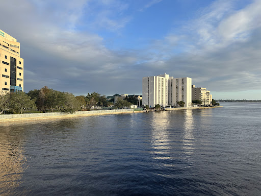 A serene view of a waterfront with modern buildings against a backdrop of clouds and clear sky.