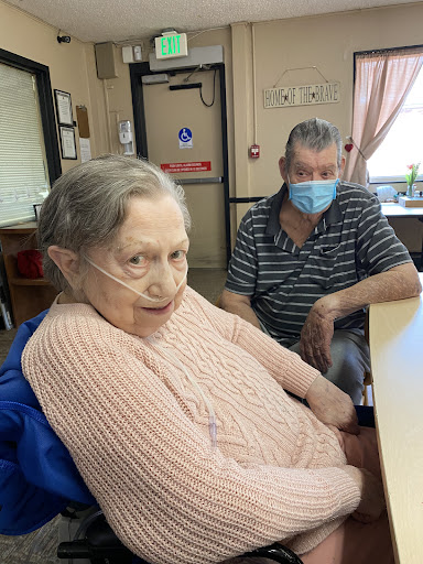 An elderly woman and a man wearing masks enjoy each other's company in a care facility.