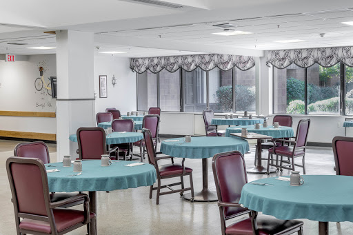 An empty dining area featuring tables with blue tablecloths and red chairs, illuminated by natural light from large windows.