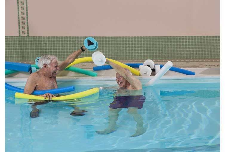 Two seniors enjoying water aerobics with pool noodles in a swimming pool.