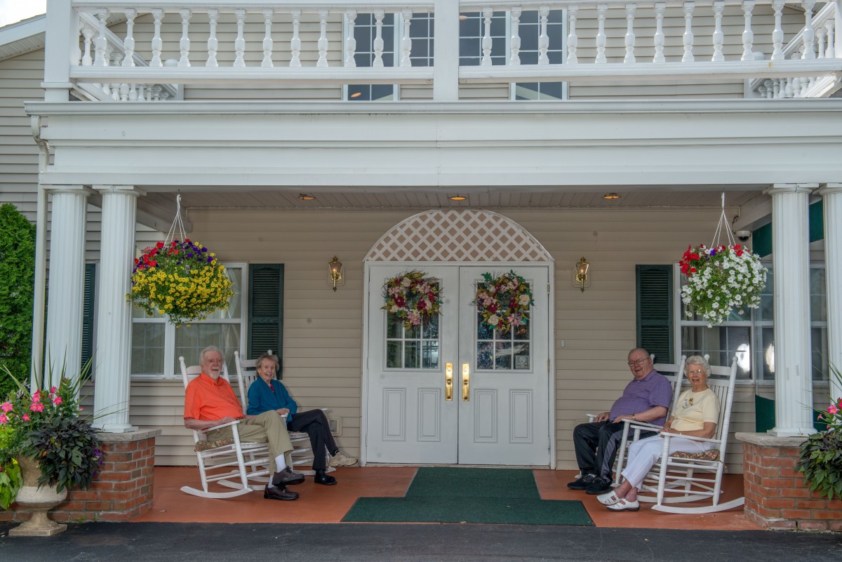 A group of people sitting in rocking chairs on a front porch by the entrance of a building, decorated with hanging flower baskets.