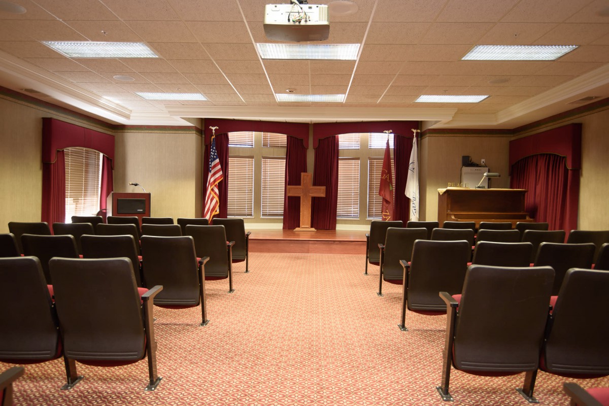 A ceremonial room with arranged seating, a podium, and flags, featuring a cross at the front.