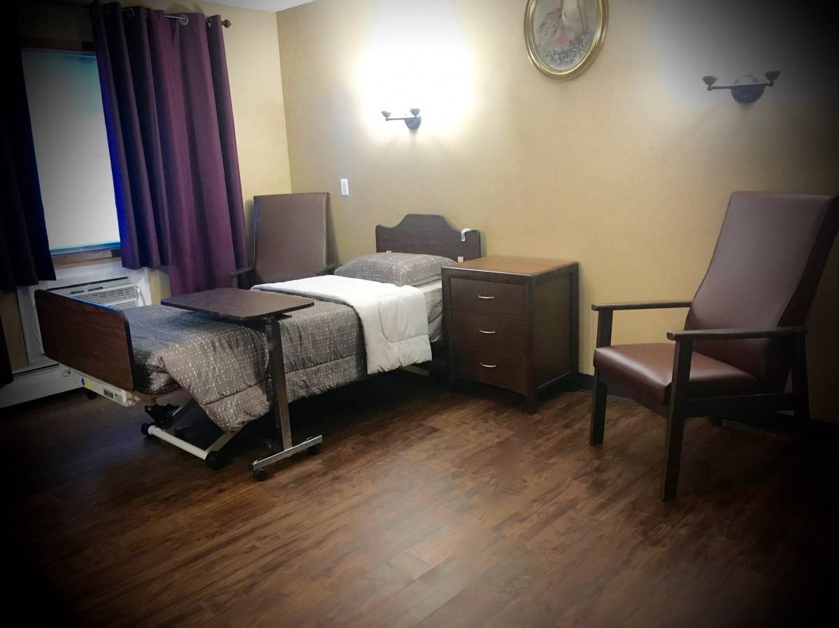 A hospital room with a bed, nightstand, and chairs on a wooden floor.