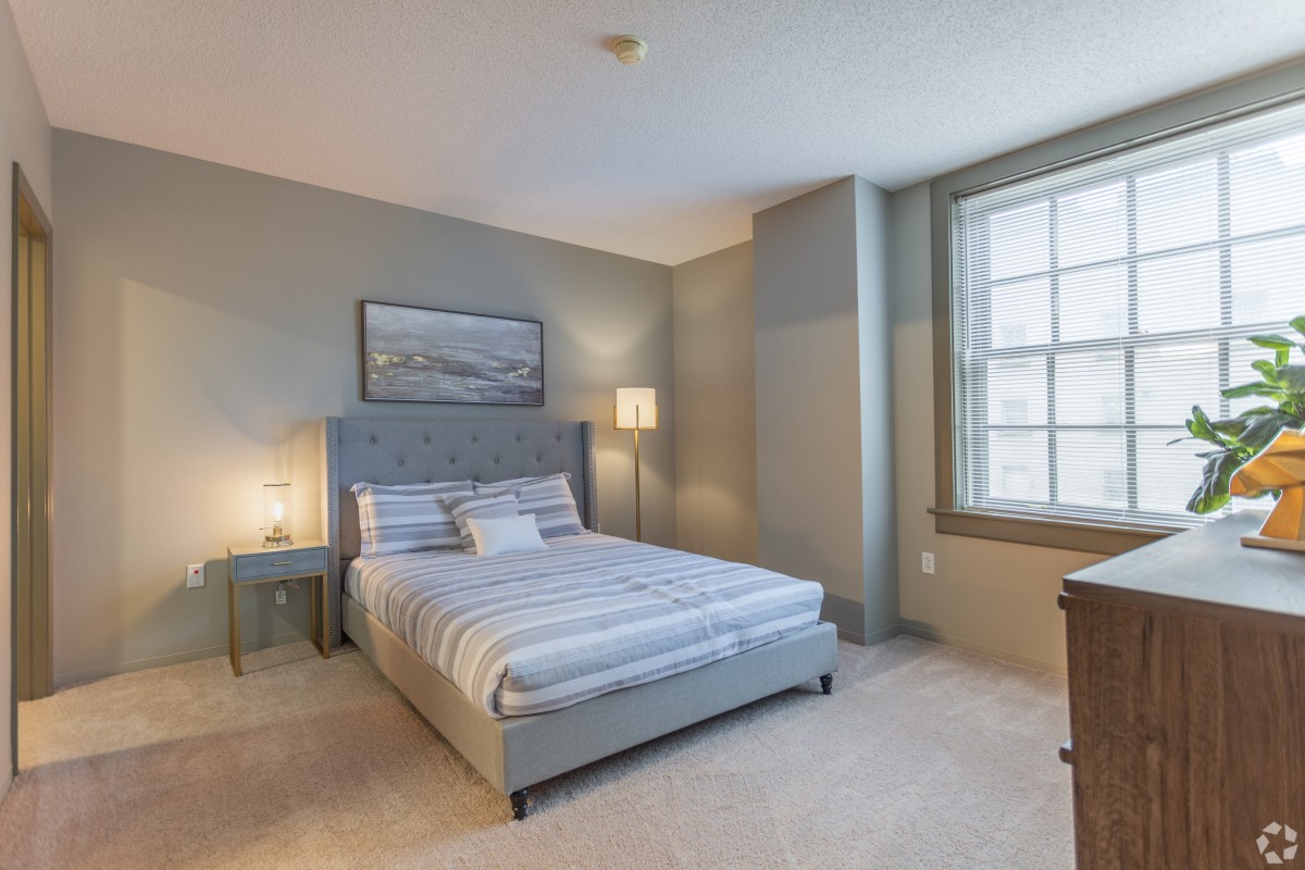A cozy bedroom interior with a neatly made bed, a bedside table with a lamp, and a large window.