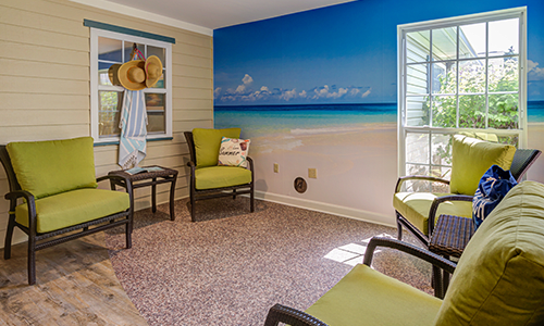 A cozy waiting room featuring a vibrant beach mural, with green cushioned chairs and natural light from a window.