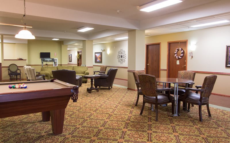 A cozy lounge area featuring a pool table, chairs, and tables on a patterned carpet.