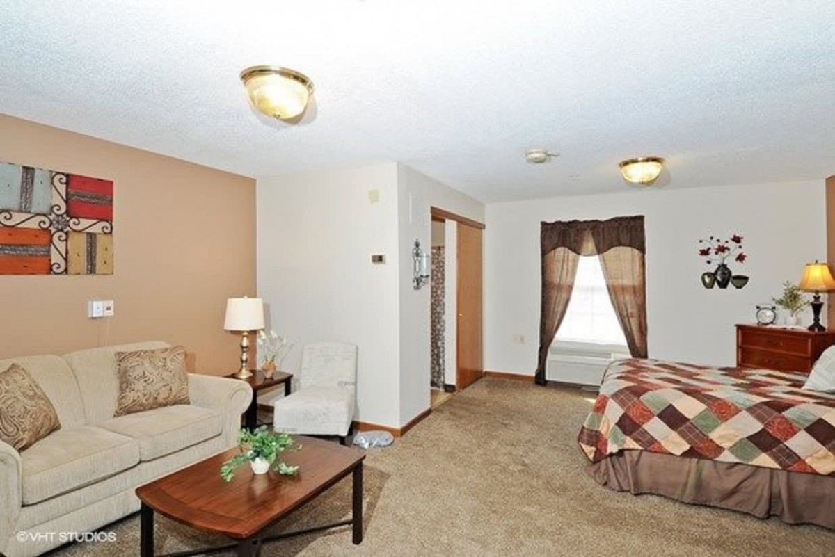 A cozy living space combining a seating area with a small sofa, coffee table, and lamp, alongside a bed with patterned covers. The room features carpeted flooring and is illuminated by ceiling lights.