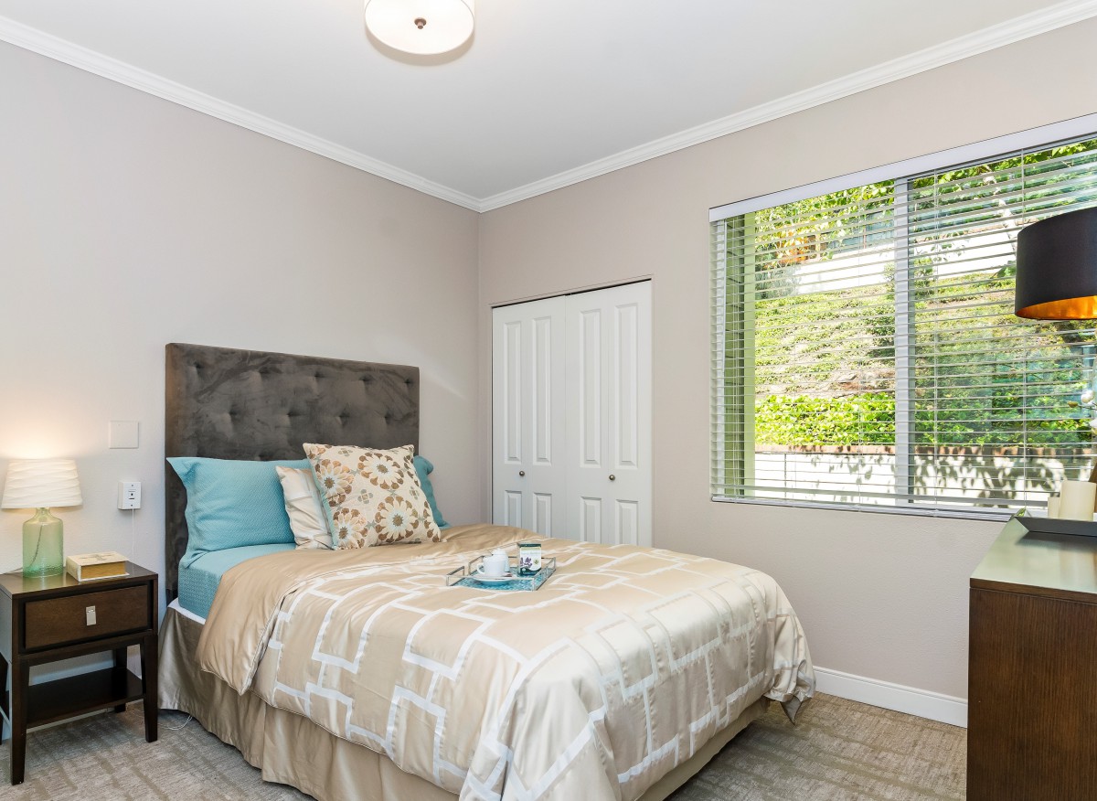 A cozy and well-lit bedroom featuring a neatly made bed with decorative pillows, a window allowing natural light, and neutral-colored walls.