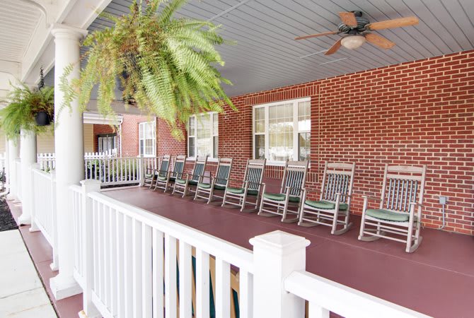 A row of rocking chairs lines the porch of a brick building, with a ceiling fan above and a potted plant hanging in the corner.