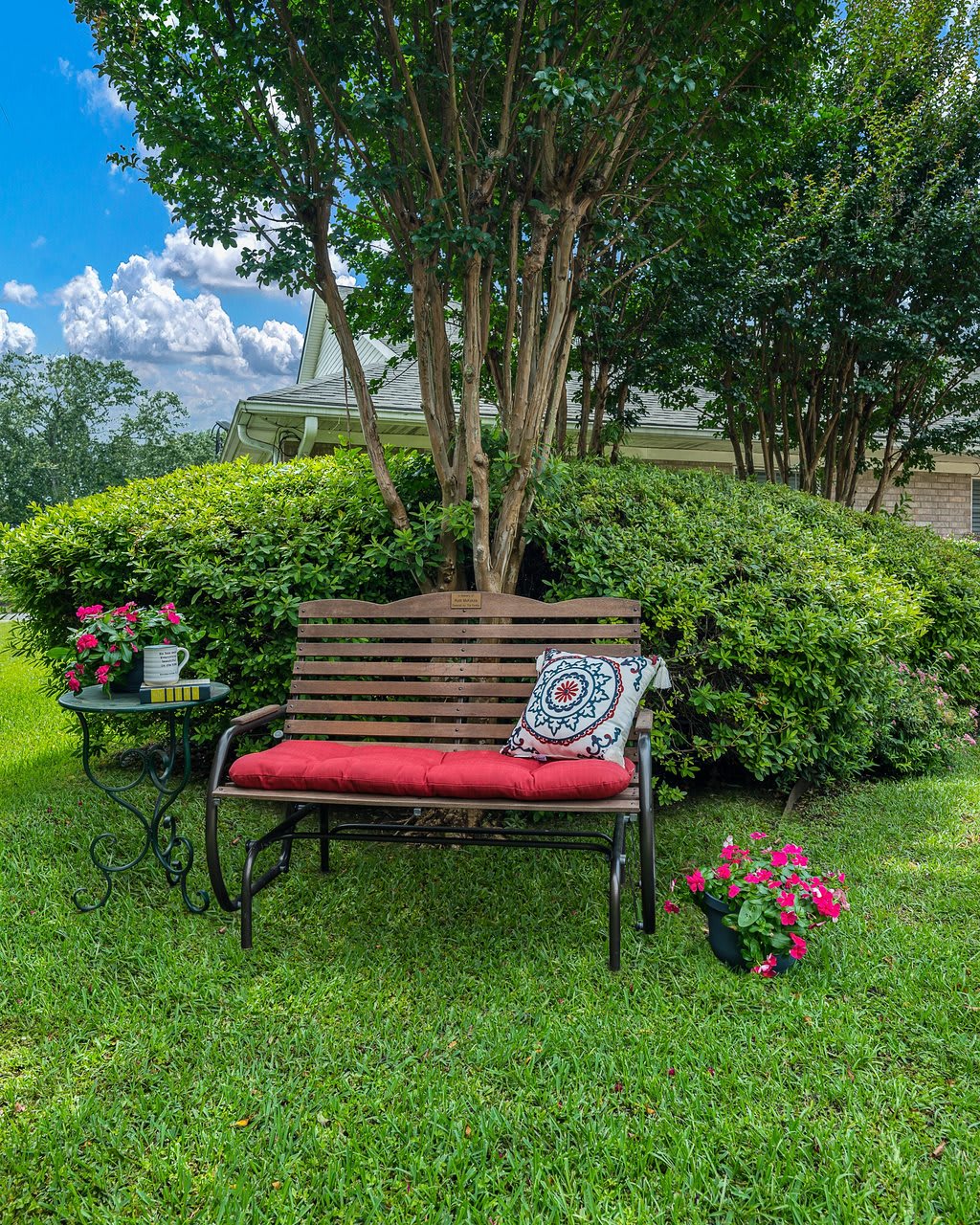 A cozy garden bench with a red cushion and decorative pillow surrounded by lush greenery and colorful flowers.