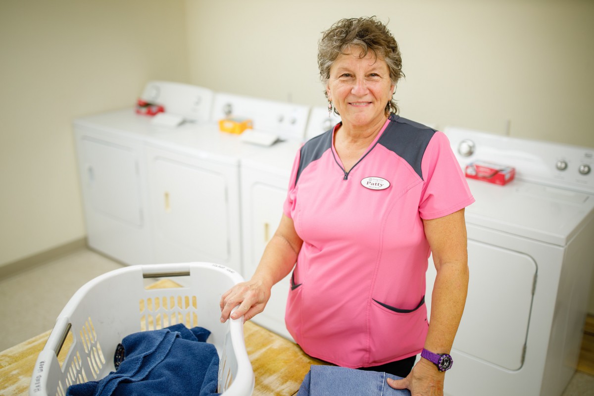 A woman in a pink uniform standing in a laundry room with washing machines and a laundry basket.