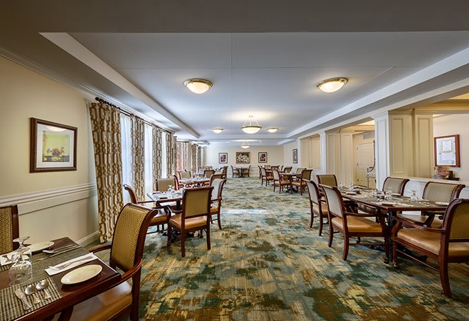 A spacious and elegantly furnished dining room with several tables and chairs, featuring patterned carpet and ample natural light from large windows.