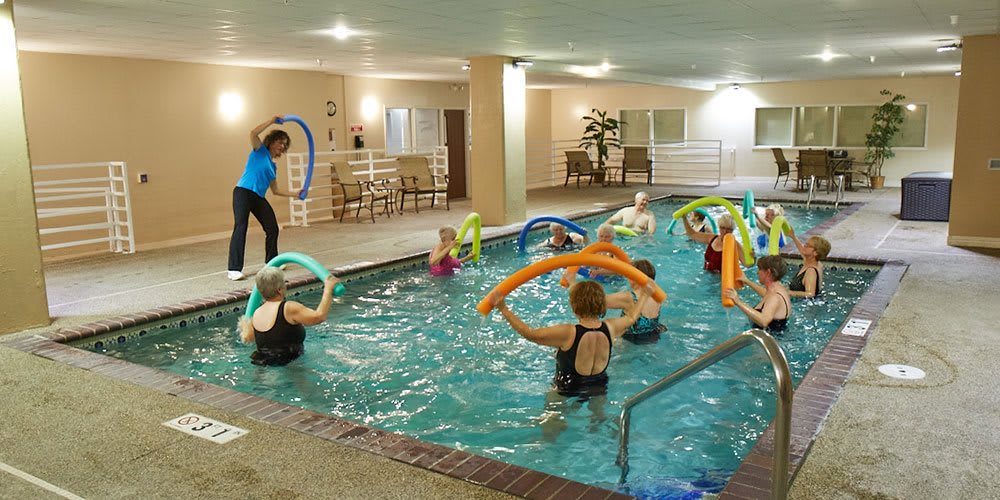 A group of people participating in an aquatic exercise class, using swim noodles in an indoor swimming pool.