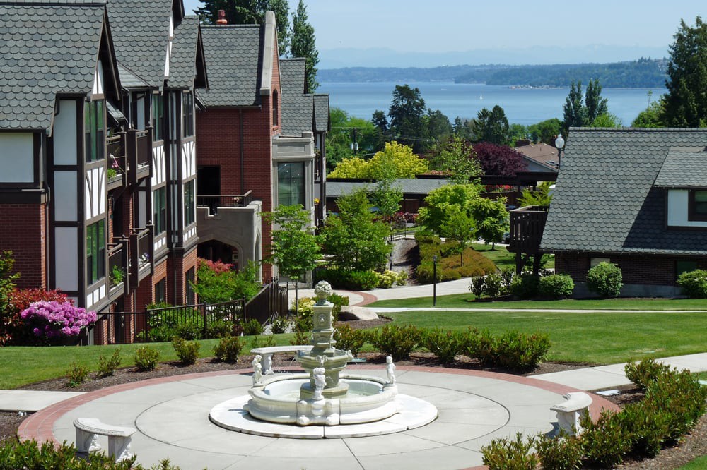A scenic neighborhood with a central fountain, surrounded by houses and greenery, overlooking a lake.