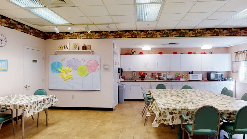 A classroom with a kitchen area, featuring a whiteboard, tables, chairs, and cabinets.