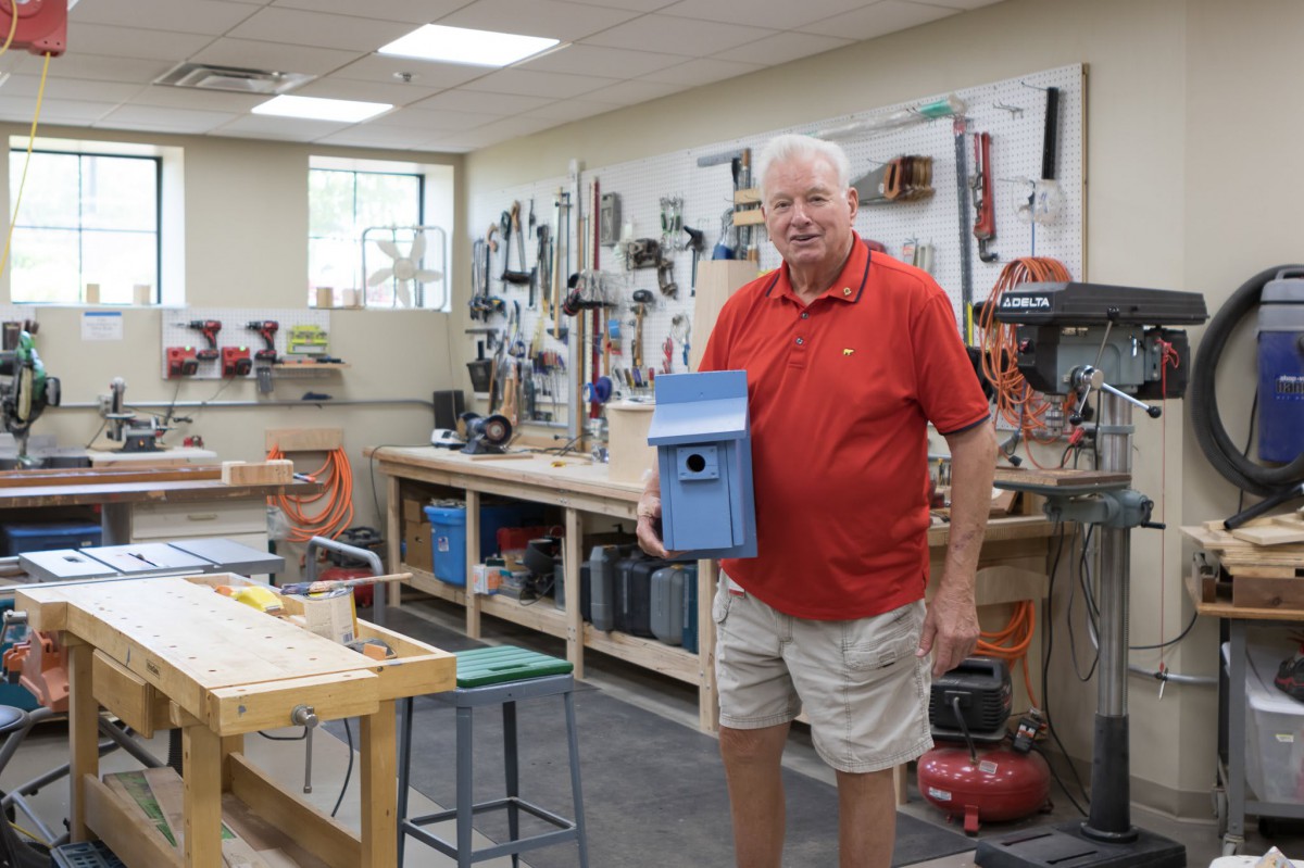A man holding a blue birdhouse in a woodworking workshop filled with various tools.
