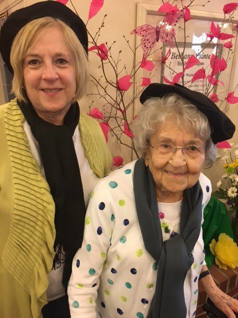 Two women smiling indoors, wearing scarves and hats, with flowers in the background.