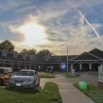 A peaceful sunset over a building with cars parked and a sidewalk in the foreground.
