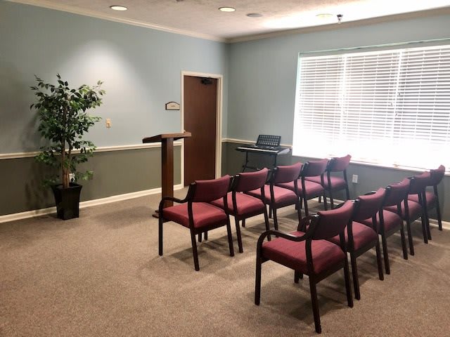 An empty room with rows of chairs facing a podium, featuring a carpeted floor, indoor plant, and window blinds.