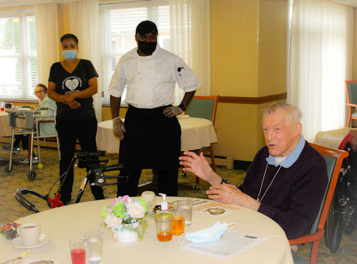 An elderly man engages in conversation at a dining table with staff members present.