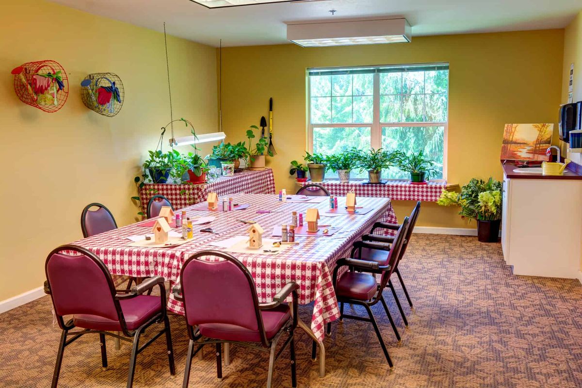 A cozy dining area with a large table adorned with a checkered tablecloth, surrounded by chairs and decorated with numerous potted plants near the window.