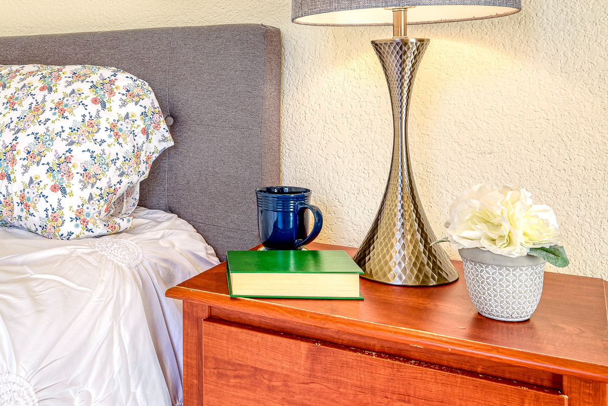 A cozy bedroom scene with a nightstand featuring a book, mug, and elegant lamp.