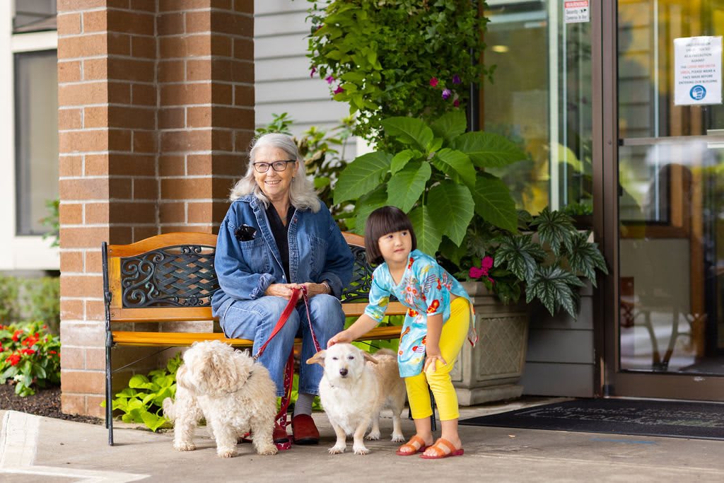 A woman and a child sitting on a bench with two fluffy dogs, surrounded by greenery.