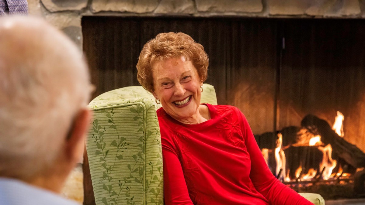 A woman in a red shirt smiling while sitting indoors near a fireplace.