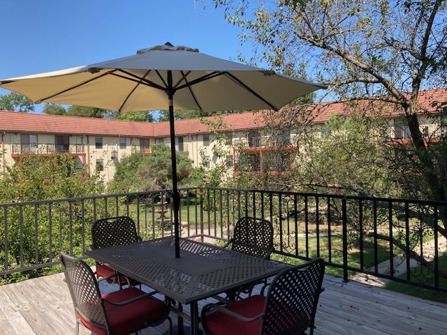 A sunny outdoor patio with a table, chairs, and an umbrella overlooking an apartment complex.