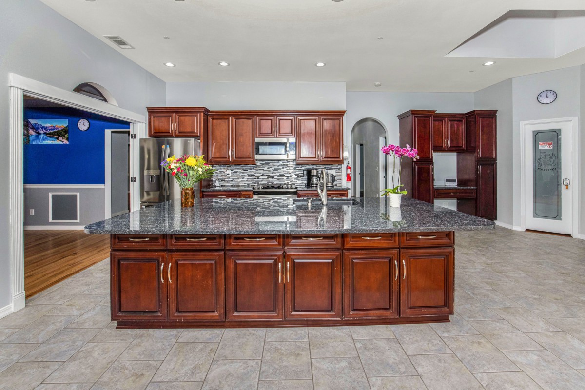 A spacious kitchen featuring wood cabinets, a large central island with a granite countertop, stainless steel appliances, and decorative flowers.