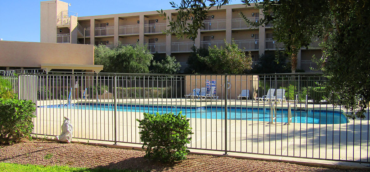 Outdoor pool area of a hotel with surrounding fence and nearby trees.