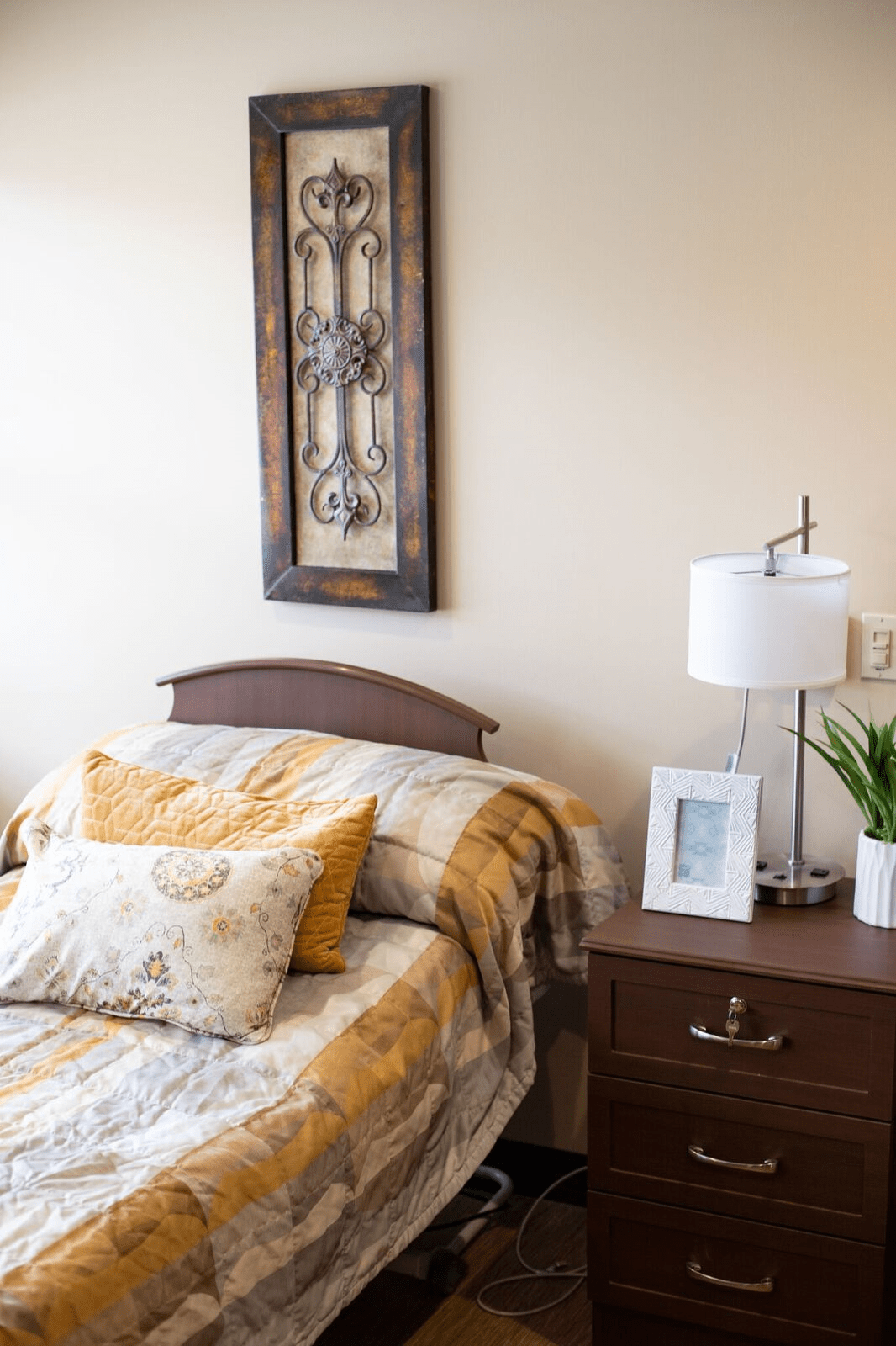 A cozy bedroom featuring a neatly made bed with patterned cushions, a wooden nightstand with a lamp, framed pictures, and a decorative plant.