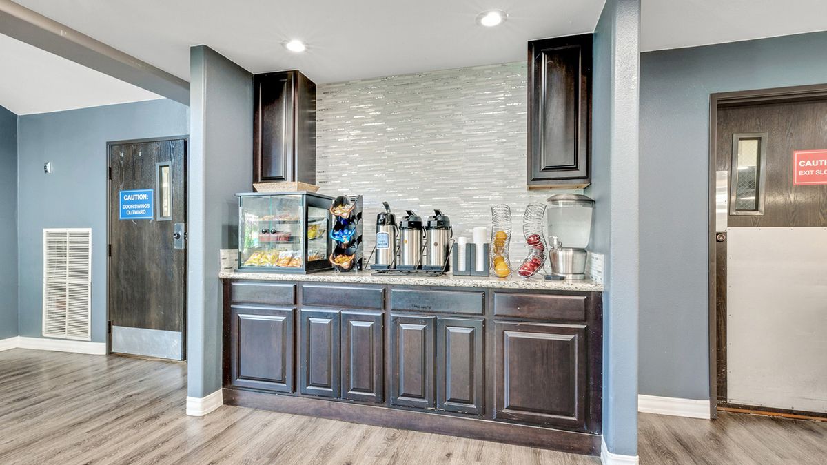 A modern coffee station with beverage dispensers and a snack display, featuring dark kitchen cabinets and wooden flooring.