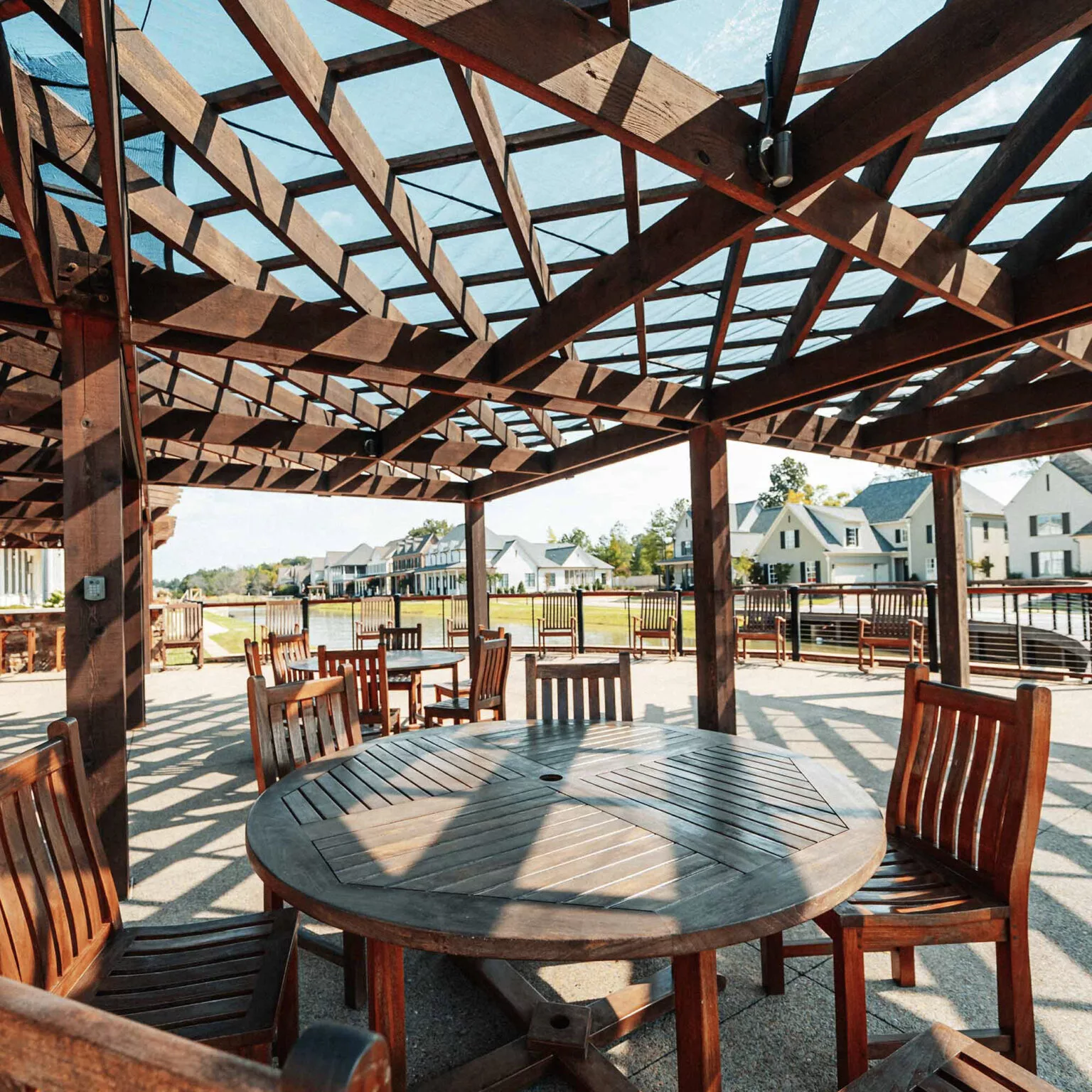 A sunny patio with wooden tables and chairs under a pergola, casting intricate shadows.