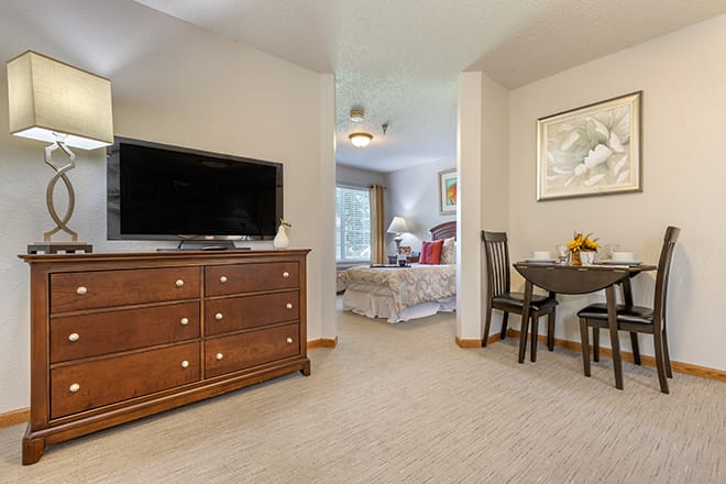 A cozy room featuring a TV on a dresser, with a view of a bedroom area and a small dining table.