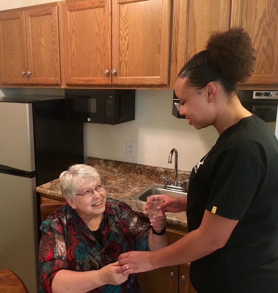 A caretaker assists an elderly woman in the kitchen, sharing a moment of connection.