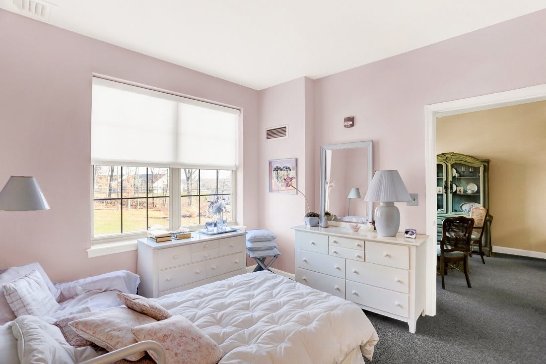 A cozy bedroom with pastel walls, featuring white furniture and a large window allowing natural light to fill the room.