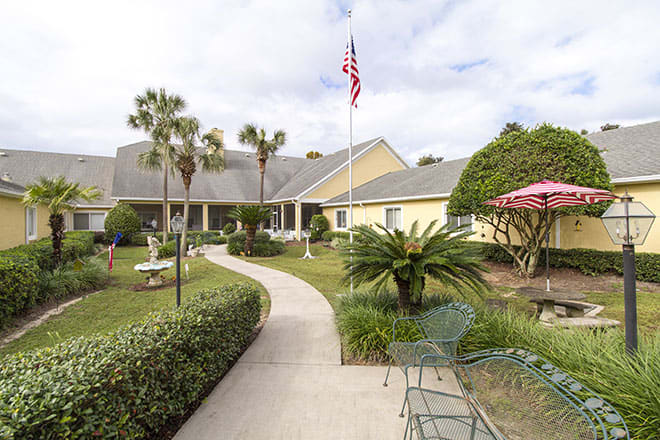 A picturesque garden pathway leads to a building, adorned with palm trees and outdoor seating, under an American flag.