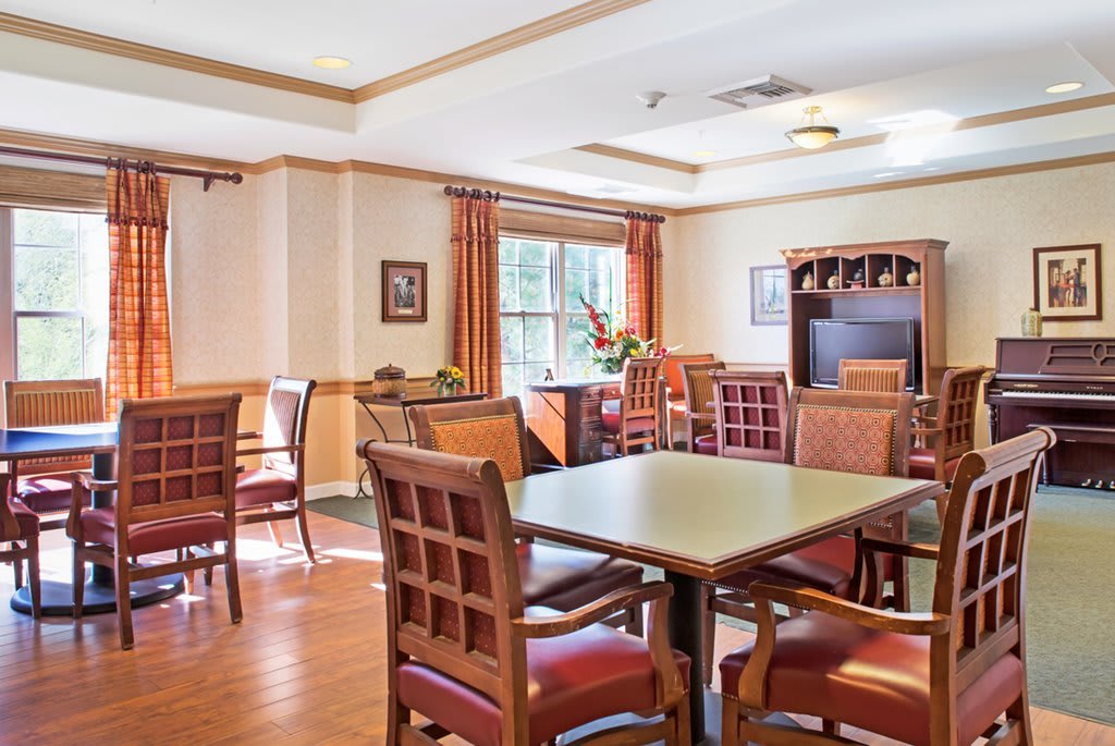 A well-lit dining room with wooden tables and chairs, featuring a large window with curtains.