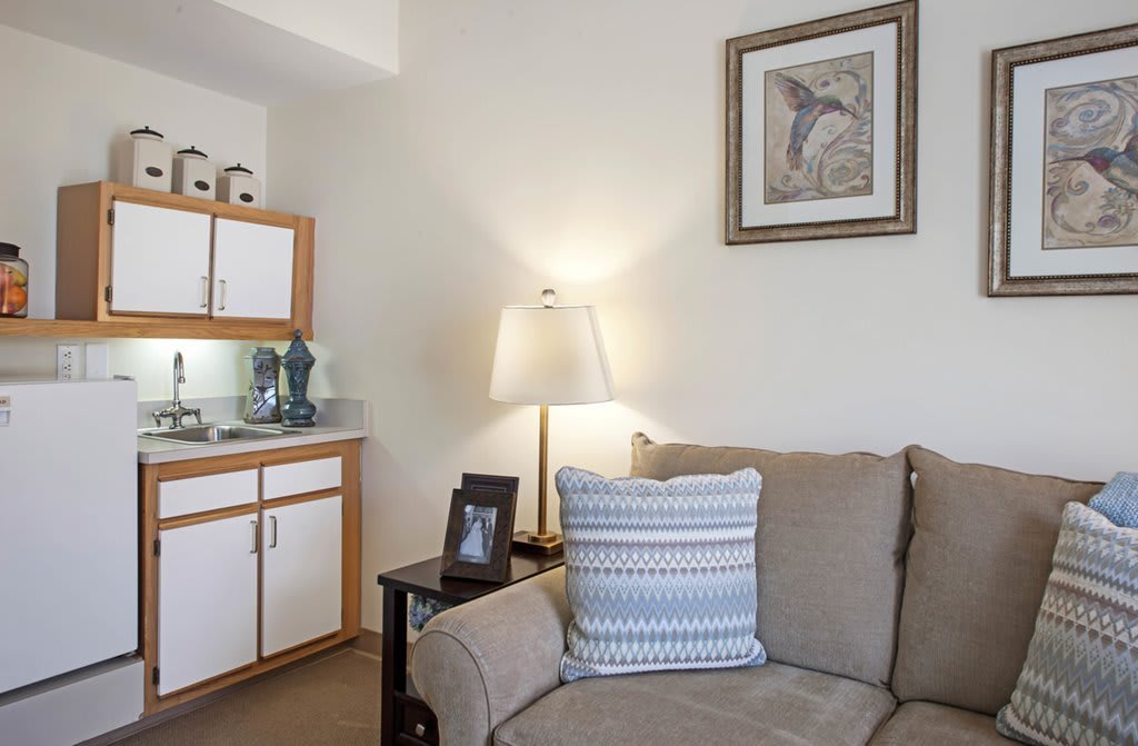 A cozy living room corner featuring a beige couch with a striped pillow, illuminated by a lamp on a side table, and adorned with decorative art on the wall.