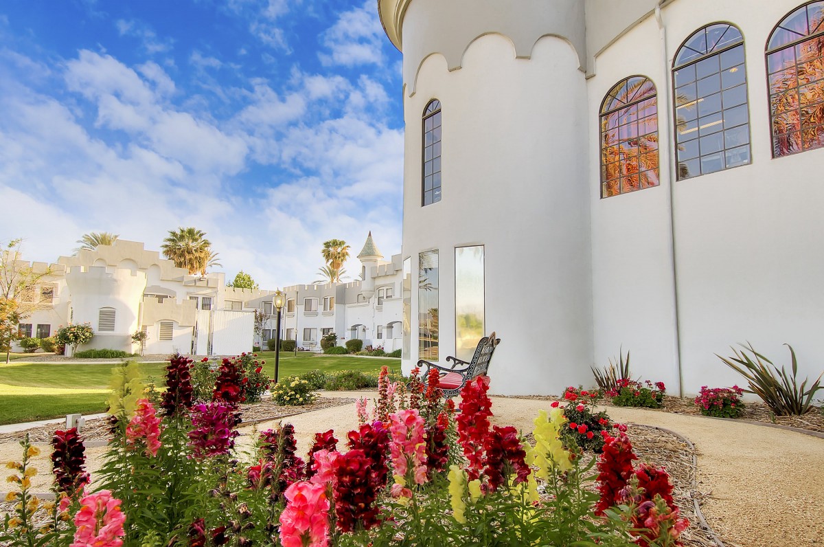A picturesque garden with vibrant flowers in front of a white building under a partly cloudy blue sky.