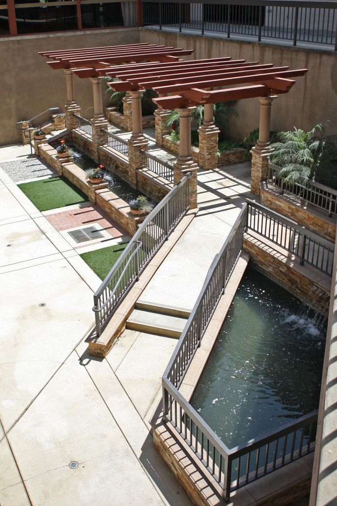 A scenic outdoor area with a wooden bridge over a water feature, surrounded by landscaped gardens and architectural elements.