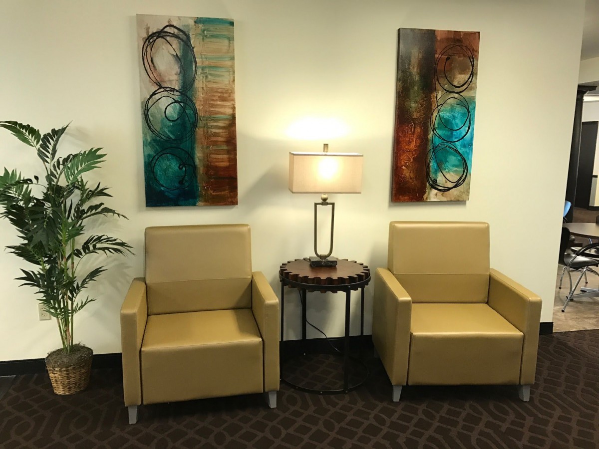 A cozy waiting area with two beige chairs, abstract art on the walls, and a lamp on a small round table.